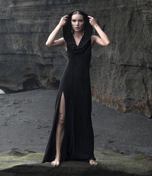 A model stands barefoot on a dark volcanic beach wearing a long black dress with a high slit and a draped hood, set against a dark rocky cliff background.