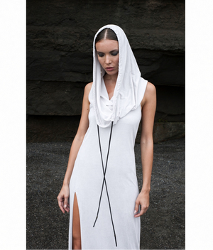 A woman wearing a white sleeveless maxi dress with a high slit and a draped hooded cowl neck tied with black leather cords, standing against a dark textured stone wall.